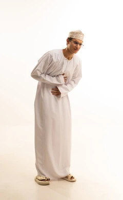 Request for help to go to the doctor, indigestion and feeling of heartburn, portrait of a young Arab Gulf Omani man wearing a dishdasha and kumma and medical glasses holding his stomach feeling pain, sensation of cramps and fatigue and exhaustion, need for medication and setting a treatment plan, full body image, white background.