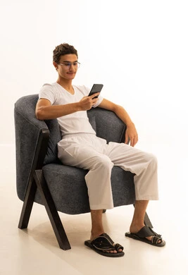 الدردشة مع الأصدقاء والأقارب عبر الجوال، ايماءات الفرح والسعادة، بورتريه لرجل عربي خليجي سعودي يرتدي السروال والفانيله البيضاء يجلس على كرسي مريح يمسك بيده الهاتف المحمول، قضاء وقت ممتع في المنزل، استخدام جهاز تقني حديث ومتطور، خلفية بيضاء