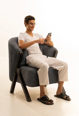 بورتريه لرجل عربي خليجي سعودي يرتدي السروال والفانيله البيضاء يجلس على كرسي مريح يمسك بيده الهاتف المحمول، قضاء وقت ممتع في المنزل، استخدام جهاز تقني حديث ومتطور، الدردشة مع الأصدقاء والأقارب عبر الجوال، ايماءات الفرح والسعادة، خلفية بيضاء