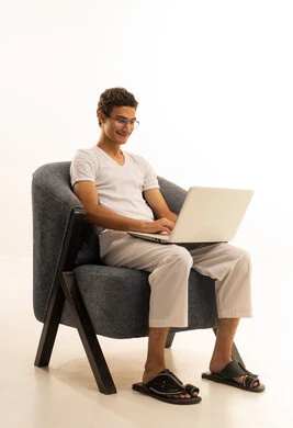 A portrait of a Saudi Arabian Gulf man wearing white pants and a white t-shirt sitting on a comfortable chair using a laptop, integrating technology with work, completing tasks required via the laptop, Saudi professions and jobs, remote business management, using modern and advanced devices, white background.