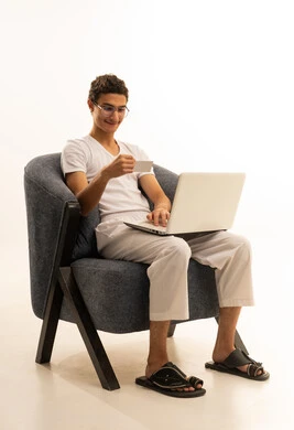 سهولة الشراء والدفع عن طريق بطاقة الإئتمان البنكية، إمكانية تسديد الفواتير بالبطاقات الإلكترونية, بورتريه لرجل عربي خليجي سعودي يرتدي سروال وفنيله يجلس على كرسي مريح ويمسك بيده بطاقة بيضاء ويعمل على الحاسوب المحمول، خلفية بيضاء