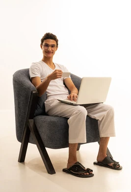 The possibility of paying bills with electronic cards, the ease of purchasing and paying with a bank credit card, a portrait of an Arab Gulf Saudi man wearing pants and a t-shirt sitting on a comfortable chair holding a white card and working on a laptop, white background.
