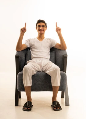 Indication of a specific location, movements that indicate this place (here), specifying the location of a specific element, a portrait of a Saudi Arabian Gulf man wearing white pants and a white shirt sitting on a comfortable chair pointing to something, white background.
