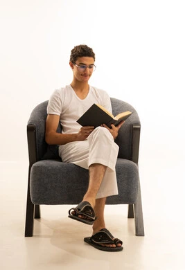 بورترية لرجل عربي خليجي سعودي يرتدي السروال والفانيله البيضاء يجلس على كرسي  مريح يمسك بيده كتاب تثقيفي ، الاستمتاع بقراءة الروايات والقصص، قراءة الكتب بتمعن، ثقافة التعلم الذاتي ،  خلفية بيضاء