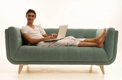 استخدام الأجهزة الحديثة والمتطورة، ادارة الأعمال عن بعد، بورتريه لرجل عربي خليجي سعودي يرتدي السروال والفانيله البيضاء يجلس على اريكة مريحة يقوم باستخدام الحاسوب المحمول، دمج التكنولوجيا بالعمل، إنجاز المهام المطلوبة عبر اللابتوب، مهن ووظائف سعودية ، خلفية بيضاء