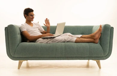 اجراء بث مباشر ، استخدام الأجهزة الحديثة والمتطورة، بورتريه لرجل عربي خليجي سعودي يرتدي السروال والفانيله البيضاء يجلس على اريكة مريحة يقوم باستخدام الحاسوب المحمول، دمج التكنولوجيا بالعمل، إنجاز المهام المطلوبة عبر اللابتوب، مهن ووظائف سعودية ، خلفية بيضاء