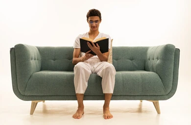 الاستمتاع بقراءة الروايات والقصص،  قراءة الكتب بتمعن، ثقافة التعلم الذاتي ، بورترية لرجل عربي خليجي سعودي يرتدي السروال والفانيله البيضاء يجلس على اريكة مريحة يمسك بيده كتاب تثقيفي ، خلفية بيضاء