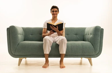 بورترية لرجل عربي خليجي سعودي يرتدي السروال والفانيله البيضاء يجلس على اريكة مريحة يمسك بيده كتاب تثقيفي ، الاستمتاع بقراءة الروايات والقصص، قراءة الكتب بتمعن، ثقافة التعلم الذاتي ، خلفية بيضاء