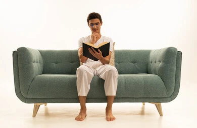 ثقافة التعلم الذاتي ، بقراءة الكتب بتمعن، ورترية لرجل عربي خليجي سعودي يرتدي السروال والفانيله البيضاء يجلس على اريكة مريحة يمسك بيده كتاب تثقيفي ، الاستمتاع بقراءة الروايات والقصص، خلفية بيضاء