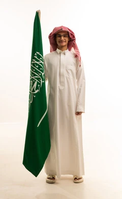 Flag Day is March 11, the celebration of the National Day on the 8th of Rabi' al-Awwal, pride and honor in belonging to the Kingdom, the anniversary of the Saudi National Day, a portrait of a smiling Arab Saudi Gulf man wearing traditional attire holding the Saudi flag in his hand, white background.