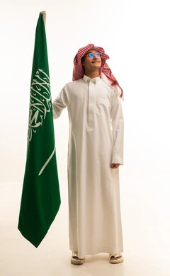 يوم العلم 11مارس، الاحتفال باليوم الوطني 8 ربيع الأول, الفخر و الاعتزاز بالانتماء للمملكة, ذكرى العيد الوطني السعودي, بورتريه لرجل عربي سعودي خليجي مبتسم يرتدي الثوب التقليدي يحمل بيده علم السعودية , خلفية بيضاء