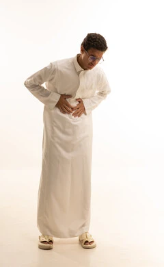 طلب المساعده للذهاب الى الطبيب، عسر الهضم والشعور بحرقة في المعدة، بورتريه لشاب عربي سعودي خليجي يرتدي الزي التقليدي يمسك بطنه شعورا بالألم، الإحساس بالمغص و التعب والإرهاق، الحاجة للدواء ووضع خطة علاجية، صورة الجسم بالكامل، خلفية بيضاء