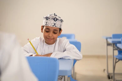 أداء التمارين الدراسية، الإستعداد للعام الدراسي الجديد، التعليم في سلطنة عمان ، العودة إلى المدارس، طالب عربي خليجي عماني يرتدي الدشداشة و الكمة يجلس في مقعده و يقوم بحل اسئلة الامتحان