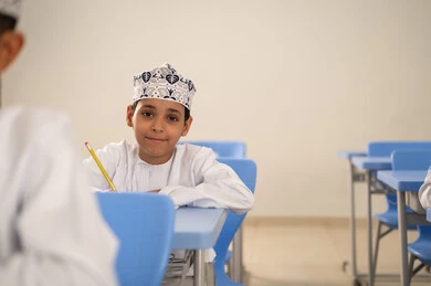 القيام بالواجبات الصفية، الإستعداد للعام الدراسي الجديد، التعليم في سلطنة عمان ، العودة إلى المدارس، طالب عربي خليجي عماني يرتدي الدشداشة و الكمة يجلس في مقعده و يقوم بحل اسئلة الامتحان