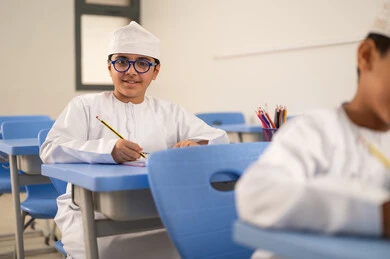 Completing academic tasks, preparing for the new school year, education in the Sultanate of Oman, back to school, an Arab Gulf Omani student wearing a dishdasha and kumma sits in his seat and solves exam questions.