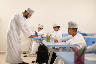 معلم عربي خليجي عماني يرتدي الدشداشة والكمة يقوم بمناقشة الدروس مع طلابه، حوارات ودية بين الاستاذ والطلاب، التربية والتعليم بالمناهج المقررة للتلاميذ، جودة التعليم في سلطنة عمان، المشاركة الفعالة للطلاب مع المدرسين