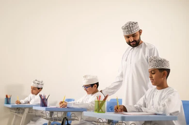 Performing school assignments, preparing for the new school year, returning to school, education in the Sultanate of Oman, a group of Arab Gulf Omani students wearing the dishdasha and kumma are sitting in their seats and doing their homework with their teacher.
