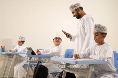 دمج التكنولوجيا في أساليب التعلم، معلم عربي خليجي عماني يقوم بتفصح الانترنت عبر الهاتف المحمول، المناهج المطورة والتعليم الالكتروني، تطور طرق التدريس في مدارس سلطنة عمان، طلاب عربيين خليجيين عمانيين يجلسون في مقاعدهم  يقومون بحل اسئله الامتحان