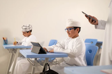 استخدام التقنيات المتطورة في الدراسة، معلم عربي خليجي عماني يقوم بتفصح الانترنت عبر الهاتف المحمول، المناهج المطورة والتعليم الالكتروني، تطور طرق التدريس في مدارس سلطنة عمان، طلاب عربيين خليجيين عمانيين يجلسون في مقاعدهم  يقومون بحل اسئله الامتحان