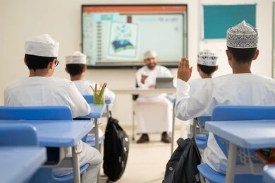 Raising the hand to request permission to answer, benefiting from modern technologies and techniques in learning and studying, developed curricula and e-learning, the evolution of teaching methods in schools in the Sultanate of Oman, two Arab Gulf Omani students sitting in their seats with an Omani teacher using a tablet in front of them.
