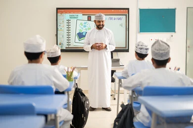 تعزيز البحث العلمي وتنمية المهارات، متابعة مجموعة من الطلاب داخل الفصل، معلم عربي خليجي عماني يرتدي الدشداشه والكمه يشرح لطلابه في بيئه مدرسيه تعليميه متطوره، نظام التعليم الحديث، جودة التعلم في المدارس الأكاديميه