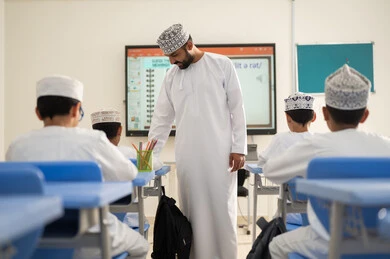 Enhancing innovation and developing scientific skills, monitoring a group of students inside the classroom, a Gulf Arab Omani teacher wearing a dishdasha and kumma explaining to his students in an advanced educational school environment, the modern education system, the quality of learning in academic schools.