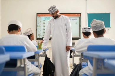 تنمية المهارات وتشجيع الابتكار العلمي، متابعة مجموعة من الطلاب داخل الفصل، معلم عربي خليجي عماني يرتدي الدشداشه والكمه يشرح لطلابه في بيئه مدرسيه تعليميه متطوره، نظام التعليم الحديث، جودة التعلم في المدارس الأكاديميه