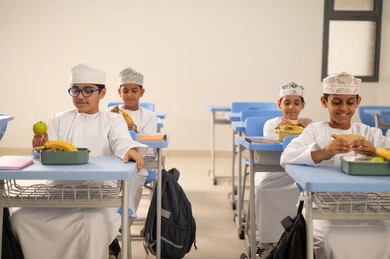 Eating nutritious meals during the break, the back-to-school season, foods rich in essential nutrients for building the body, a group of Arab Gulf Omani students wearing the dishdasha and kumma are sitting in their seats with lunch boxes in front of them.