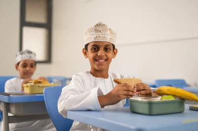 التغذية الصحية في فترات الاستراحة، موسم العودة للمدارس، الوجبات والسناكات الغنية بالعناصر الغذائية الهامة لبناء الجسم، مجموعة من الطلاب العربيين الخليجيين العمانيين يرتدون الدشداشة والكمة يجلسون في مقاعدهم وأمامهم اللانش البوكس