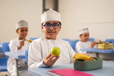 تناول وجبات صحية بين الحصص، موسم العودة للمدارس، الوجبات والسناكات الغنية بالعناصر الغذائية الهامة لبناء الجسم، مجموعة من الطلاب العربيين الخليجيين العمانيين يرتدون الدشداشة والكمة يجلسون في مقاعدهم وأمامهم اللانش البوكس