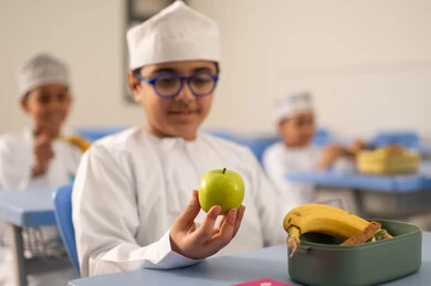 تناول وجبات مفيدة أثناء الاستراحة، موسم العودة للمدارس، الوجبات والسناكات الغنية بالعناصر الغذائية الهامة لبناء الجسم، مجموعة من الطلاب العربيين الخليجيين العمانيين يرتدون الدشداشة والكمة يجلسون في مقاعدهم وأمامهم اللانش البوكس