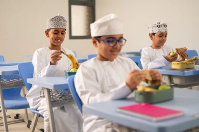 Healthy eating during the school break, back-to-school season, meals and snacks rich in essential nutrients for building the body, a group of Arab Gulf Omani students wearing the dishdasha and kumma sitting in their seats with lunch boxes in front of them.