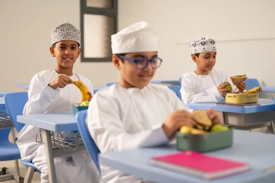 تناول الأطعمة الصحية في وقت الفسحة المدرسية، موسم العودة للمدارس، الوجبات والسناكات الغنية بالعناصر الغذائية الهامة لبناء الجسم، مجموعة من الطلاب العربيين الخليجيين العمانيين يرتدون الدشداشة والكمة يجلسون في مقاعدهم وأمامهم اللانش البوكس