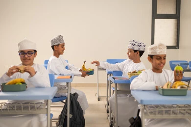Back to school season, exchanging fruit among students, eating healthy foods during school break, meals and snacks rich in essential nutrients for building the body, a group of Arab Gulf Omani students wearing the dishdasha and kumma sitting in their seats with lunch boxes in front of them.