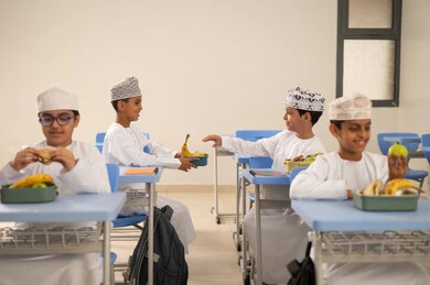 تبادل الفاكهة بين الطلاب، تناول الأطعمة الصحية في وقت الفسحة المدرسية، موسم العودة للمدارس، الوجبات والسناكات الغنية بالعناصر الغذائية الهامة لبناء الجسم، مجموعة من الطلاب العربيين الخليجيين العمانيين يرتدون الدشداشة والكمة يجلسون في مقاعدهم وأمامهم اللانش البوكس