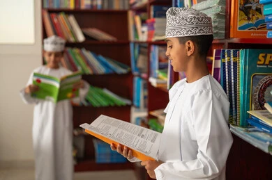 Self-education during free time, a love for exploration and knowledge, two Arab Gulf students from Oman wearing the dishdasha and kumma are reading school books, education and study in the Sultanate of Oman, developing reading skills for students, visiting the school library.