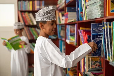 حب الاطلاع والمعرفة، طالب عربي خليجي عماني يرتدي الدشداشة و الكمة يقوم باختيار كتاب، التعليم والدراسة في سلطنة عمان ، قضاء وقت الفراغ بالتعلم والتثقف الذاتي, تنمية مهارة القراءة للطلاب, زيارة مكتبة المدرسة