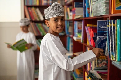 زيارة مكتبة المدرسة، حب الثقافة والاستطلاع، طالب عربي خليجي عماني يرتدي الدشداشة و الكمة يقوم باختيار كتاب، التعليم والدراسة في سلطنة عمان ، قضاء وقت الفراغ بالتعلم والتثقف الذاتي, تنمية مهارة القراءة للطلاب