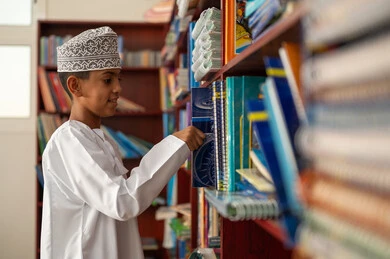 حب الثقافة والاستطلاع، طالب عربي خليجي عماني يرتدي الدشداشة و الكمة يقوم باختيار كتاب، التعليم والدراسة في سلطنة عمان ، قضاء وقت الفراغ بالتعلم والتثقف الذاتي, تنمية مهارة القراءة للطلاب, زيارة مكتبة المدرسة