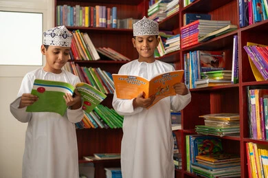 Enjoying reading books, two Arab Gulf students from Oman wearing the dishdasha and kumma stand in the school library, the concept of self-directed learning, an advanced educational school environment, academic schools in the Sultanate of Oman.