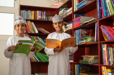 Enjoying reading books, two Arab Gulf students from Oman wearing the dishdasha and kumma stand in the school library, the concept of self-directed learning, an advanced educational school environment, academic schools in the Sultanate of Oman.