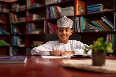 Spending free time learning and self-education, developing reading skills for students, loving culture and exploration, an Arab Gulf Omani student wearing a dishdasha and kumma is reading a book, visiting the school library, education and study in the Sultanate of Oman, and the interest in browsing useful books.