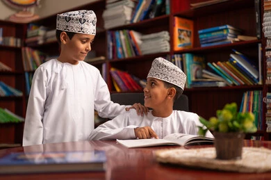 الدعم المتبادل بين الطلاب، حل التمارين التعليمية، التعليم في سلطنة عمان، الإستعداد للعام الدراسي الجديد، العودة إلى المدارس،البان عربيان خليجيان عمانيان يرتديان الدشداشة و الكمة يجلسان داخل المكتبة المدرسية يقومون بمناقشة الدروس