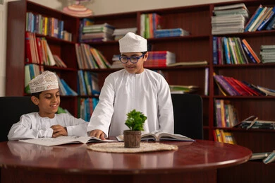 حل التمارين التعليمية، التعليم في سلطنة عمان، الإستعداد للعام الدراسي الجديد، العودة إلى المدارس، طالبان عربيان خليجيان عمانيان يرتديان الدشداشة والكمة يجلسان داخل المكتبة المدرسية يقومان بمناقشة الدروس