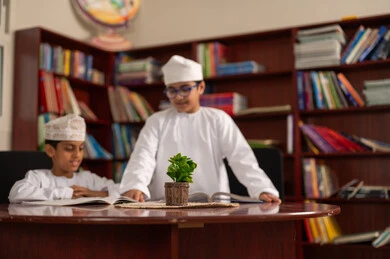 تنفيذ الواجبات الصفية، التعليم في سلطنة عمان، الإستعداد للعام الدراسي الجديد، العودة إلى المدارس، طالبان عربيان خليجيان عمانيان يرتديان الدشداشة والكمة يجلسان داخل المكتبة المدرسية يقومان بمناقشة الدروس