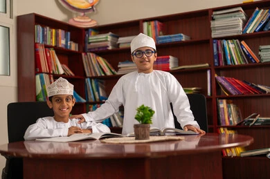 النظر للكاميرا بتعابير السعادة و السرور، التعليم في سلطنة عمان، تأدية الواجبات المدرسية، الإستعداد للعام الدراسي الجديد، العودة إلى المدارس، طالبان عربيان خليجيان عمانيان يرتديان الدشداشة والكمة يجلسان داخل المكتبة المدرسية يقومان بمناقشة الواجب