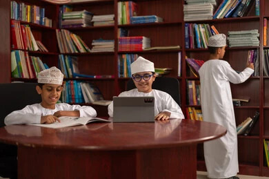 الاهتمام بالمعرفة وتنمية الفكر، تطور طرق التعليم ،المناهج المطورة والتعليم الالكتروني، الاستفادة من التقنيات والتكنولوجيا الحديثة في التعلم والدراسة،  مجموعه من الطلاب العرب الخليجيون العمانيون يرتدون الدشداشه والكمه يجلسون داخل المكتبة المدرسية، استخدام جهاز التابلت، اداء الواجبات المدرسية