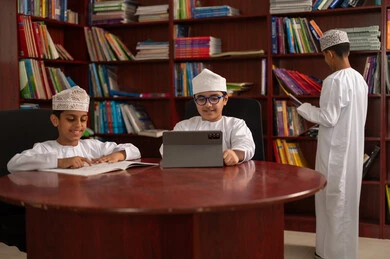 حب التعلم واستكشاف الأفكار، تطور طرق التعليم ،المناهج المطورة والتعليم الالكتروني، الاستفادة من التقنيات والتكنولوجيا الحديثة في التعلم والدراسة،  مجموعه من الطلاب العرب الخليجيون العمانيون يرتدون الدشداشه والكمه يجلسون داخل المكتبة المدرسية، استخدام جهاز التابلت، اداء الواجبات المدرسية