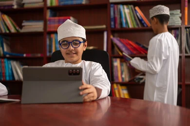 تطور طرق التعليم ،المناهج المطورة والتعليم الالكتروني، الاستفادة من التقنيات والتكنولوجيا الحديثة في التعلم والدراسة، طالبان عربيان خليجيان عمانيان يرتديان الدشداشه والكمه يجلسان في المكتبة المدرسية، استخدام جهاز التابلت، حب الثقافة والاستطلاع