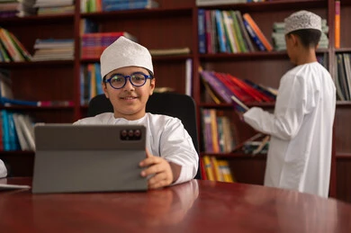 Developed curricula and e-learning, benefiting from modern technologies and techniques in learning and studying, the evolution of teaching methods, two Arab Gulf students from Oman wearing the dishdasha and kumma sitting in the school library, using a tablet device, love of culture and exploration.